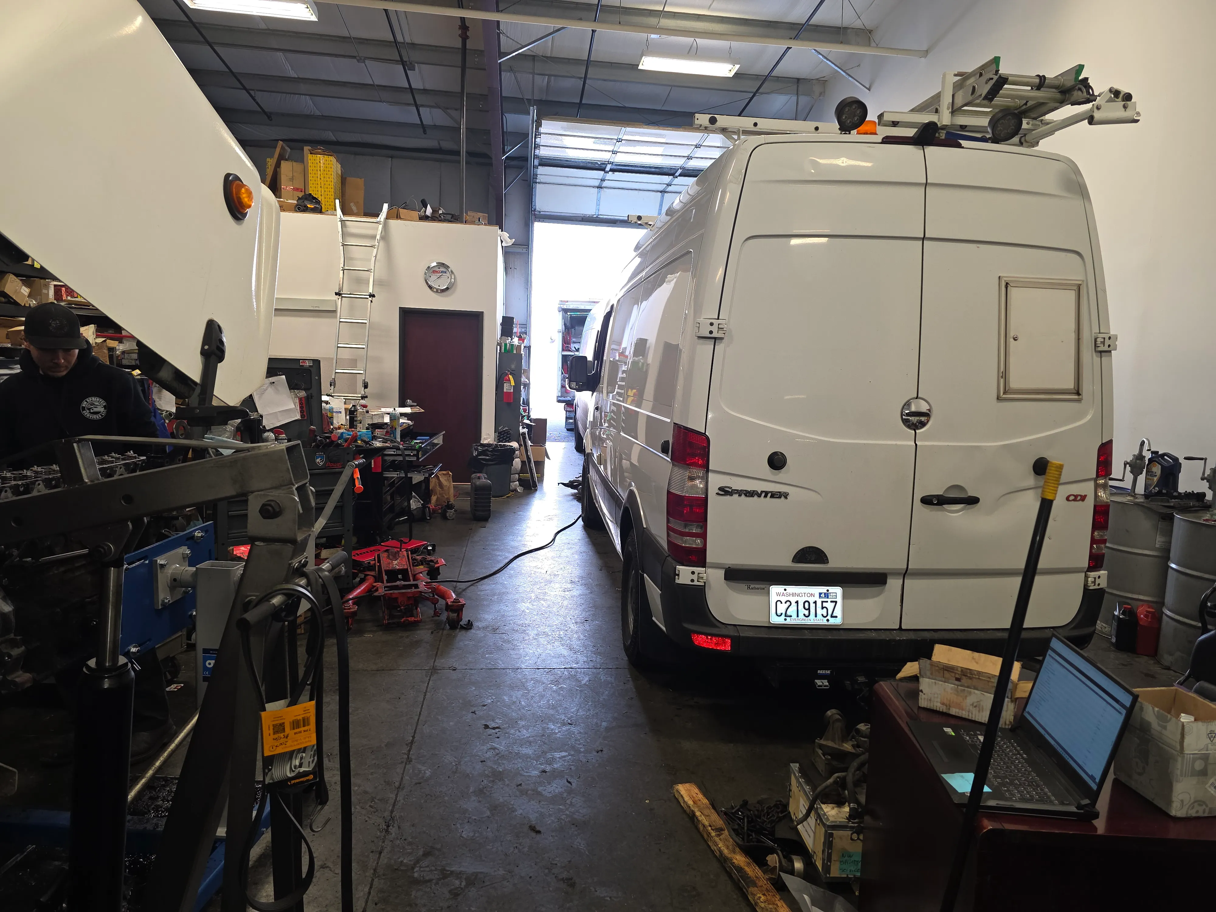 A Mercedes-Benz Sprinter being serviced on the shop floor — engine bay open, technician at work